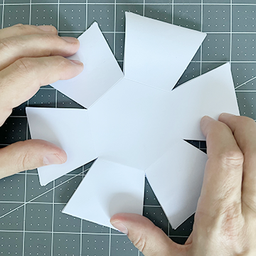 Image of two hands holding the pop-up calendar flat while the glue is drying between the tabs. Once the hands release the flat shape, the rubber band tension will pop up the calendar into a 3D shape.